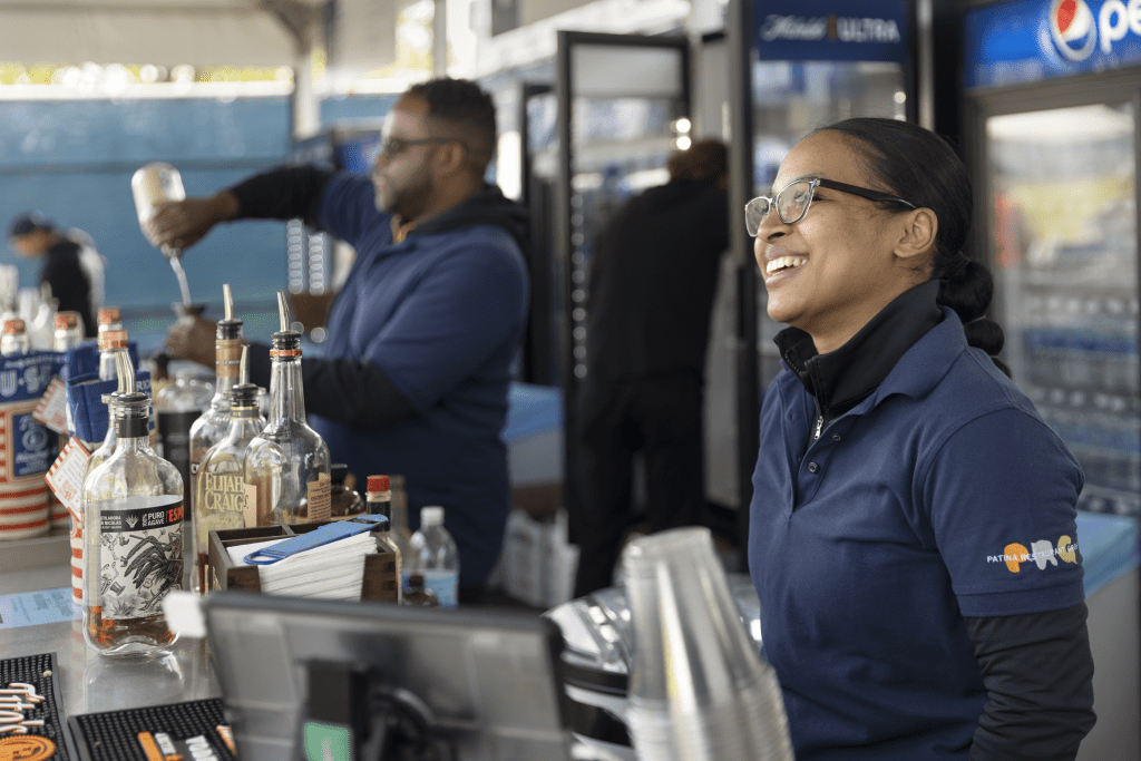 Two bartenders, one making a drink and the second smiling at guest