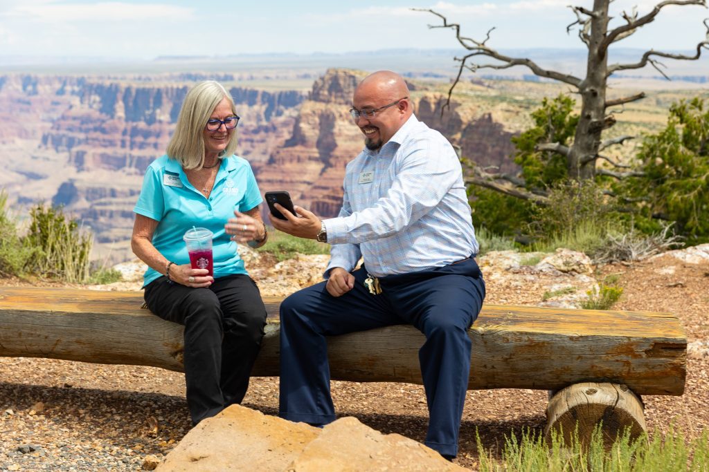 Two team members sitting with a drink looking at their phone in front of the Grand Canyon
